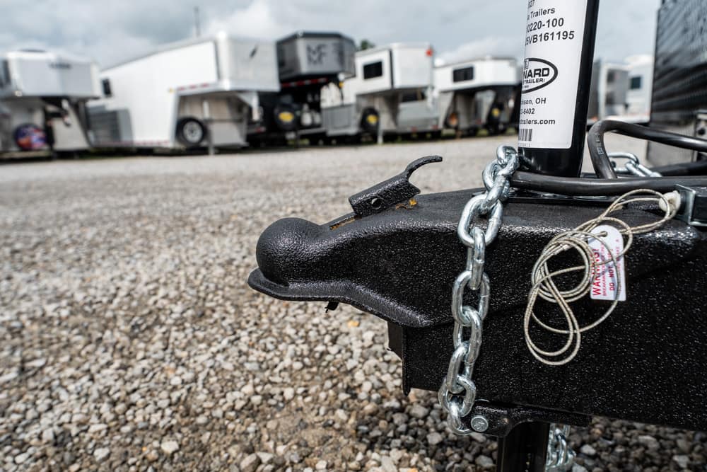 Close-up of a black trailer hitch with a steel chain, parked among several utility trailers in a gravel lot.