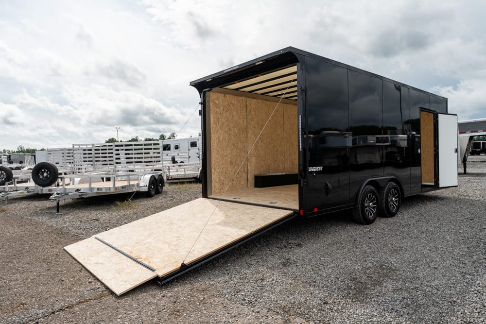 Black enclosed cargo trailer with its rear ramp lowered, exposing a plywood interior on a gravel lot with other trailers in the background.
