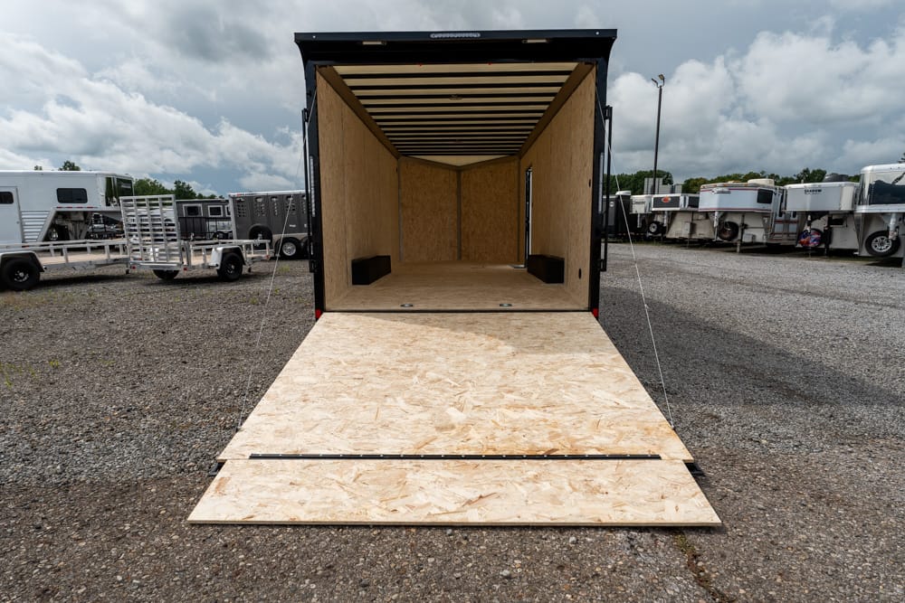 Open empty cargo trailer with plywood interior and ramp down, parked in a gravel lot with other trailers nearby.