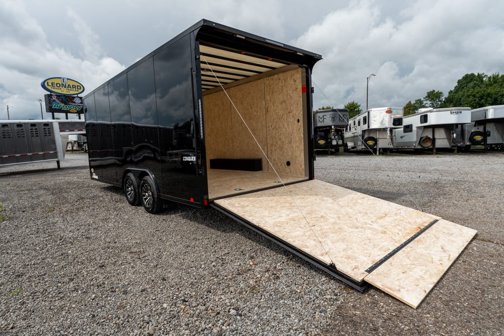 Black enclosed cargo trailer with its plywood loading ramp extended on a gravel lot, other RV trailers visible in the background at a dealership.
