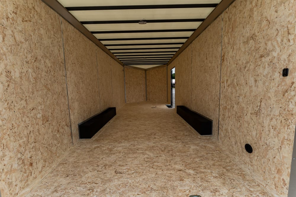 Interior of an empty plywood trailer with black wheel wells along the sides, leading to an open doorway at the far end.