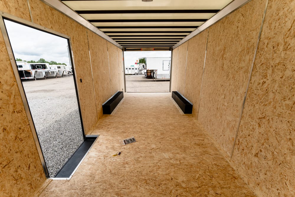 Interior of an empty plywood trailer with black storage benches along the sides and an open back door revealing trailers outside on a gravel lot.