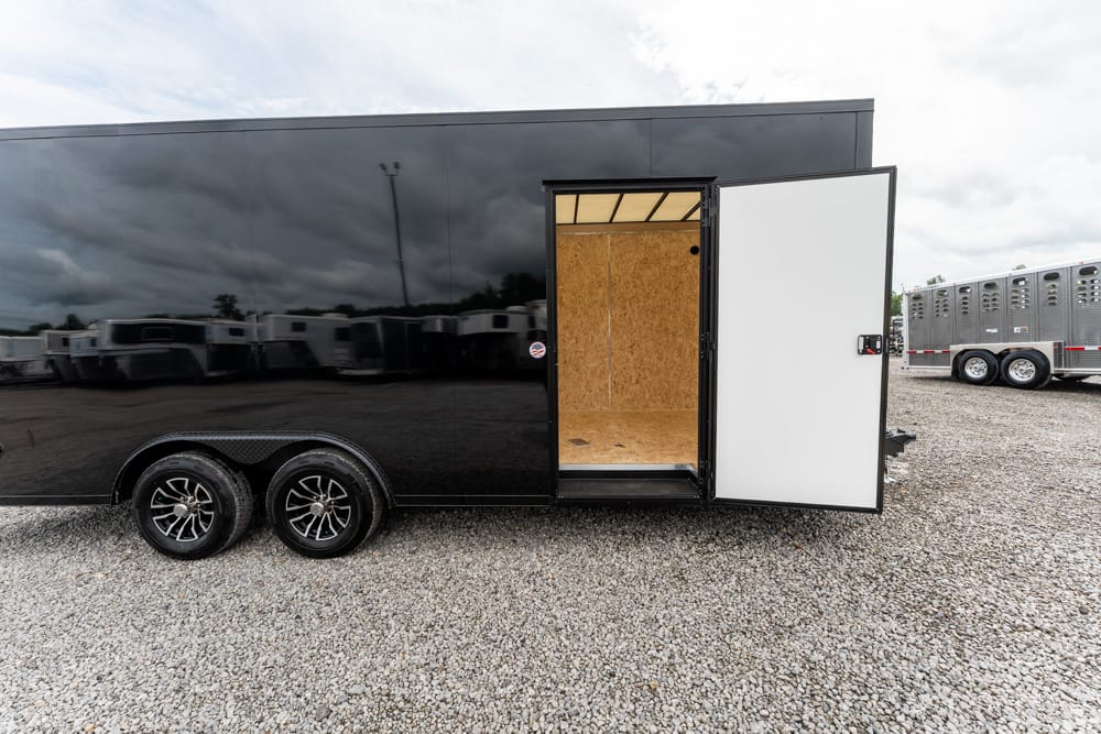 Black utility trailer with side door open, showing unfinished plywood interior and gravel ground outside.