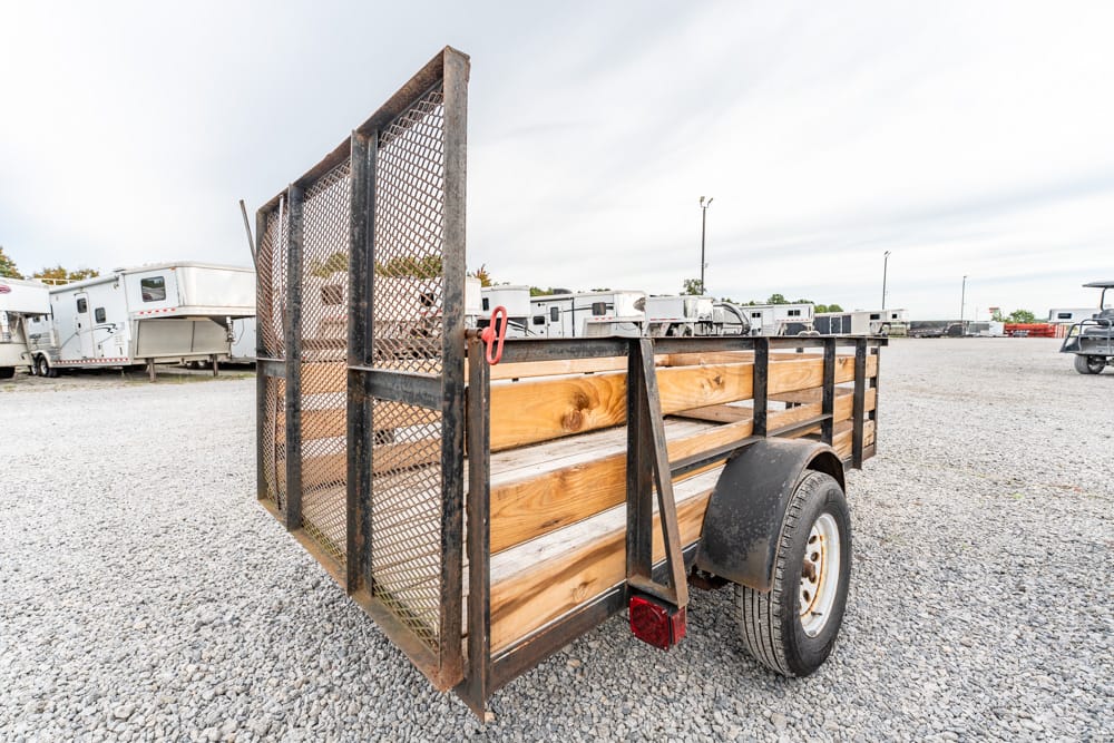 Trailer with wooden side rails and a mesh gate sits on a gravel lot, with other trailers in the background.