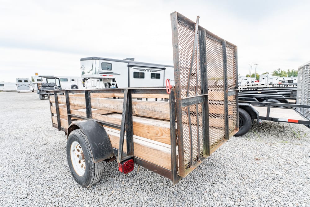 A metal-and-wood utility trailer with a mesh rear ramp parked on a gravel lot, with other trailers in the background.