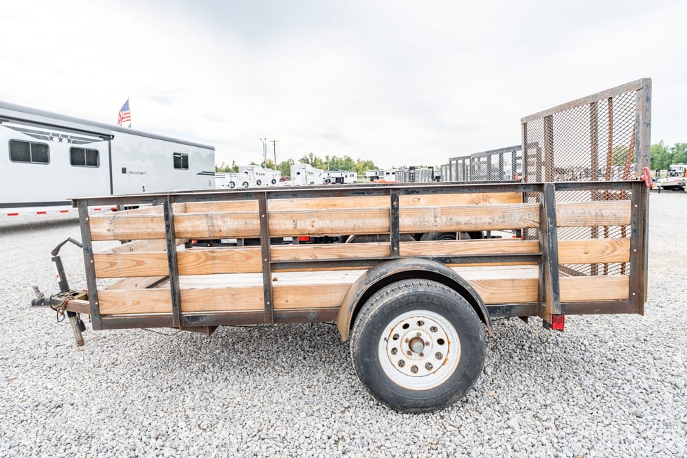 A wooden-flatbed trailer with metal frame parked on a gravel lot, hitch at the front and a single axle tire.