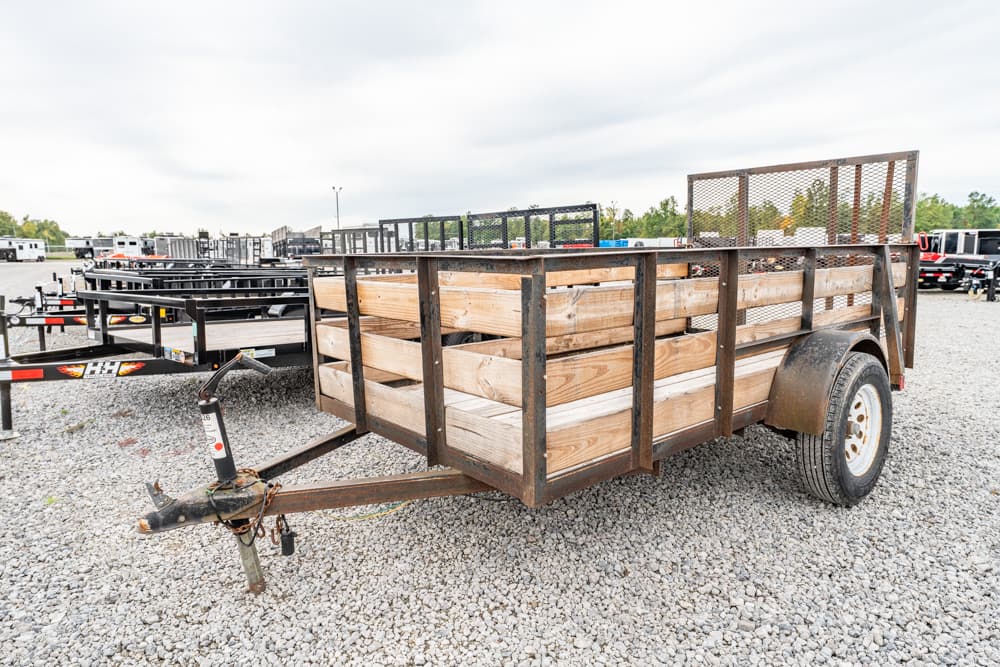 Wooden-sided utility trailer on a gravel lot, ready to haul; single-axle with a jack and safety chains.