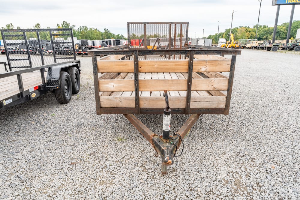 Front view of a rusted utility trailer with wooden side rails and a metal frame on a gravel lot.