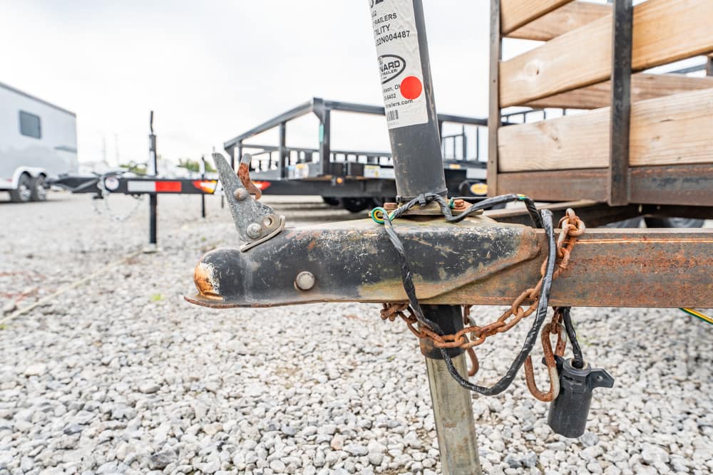 Close-up of a rusted trailer hitch with chains and a padlock on a gravel lot, background shows a wooden trailer bed and equipment. (informative)
