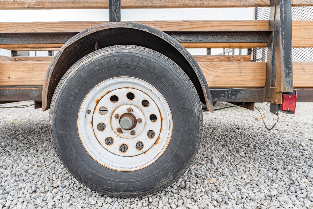 Trailer wheel with white rim and rust, attached to a weathered wooden trailer on gravel surface.