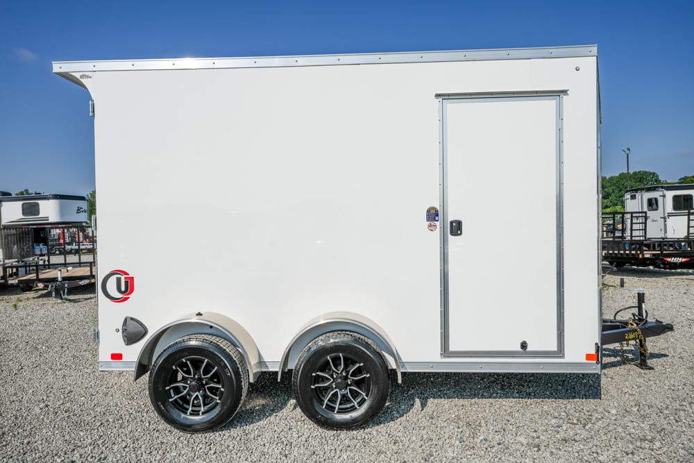 White enclosed cargo trailer with dual axles and a side entry door on a gravel lot, hitch visible at the front edge of the frame.