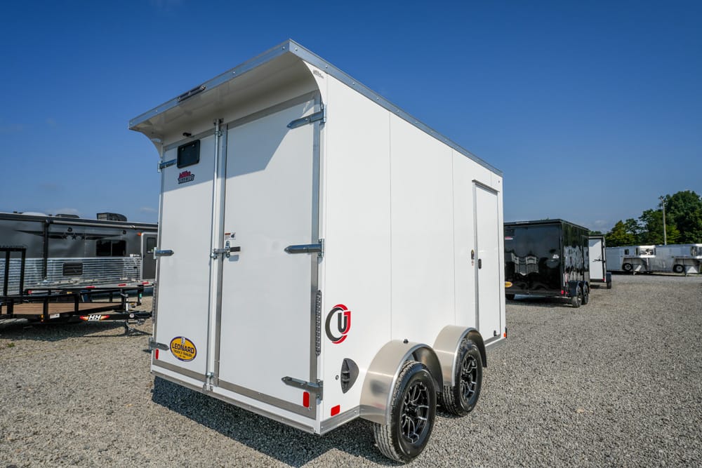 Rear three-quarter view of a white enclosed cargo trailer with dual axles parked on a gravel lot under a blue sky.