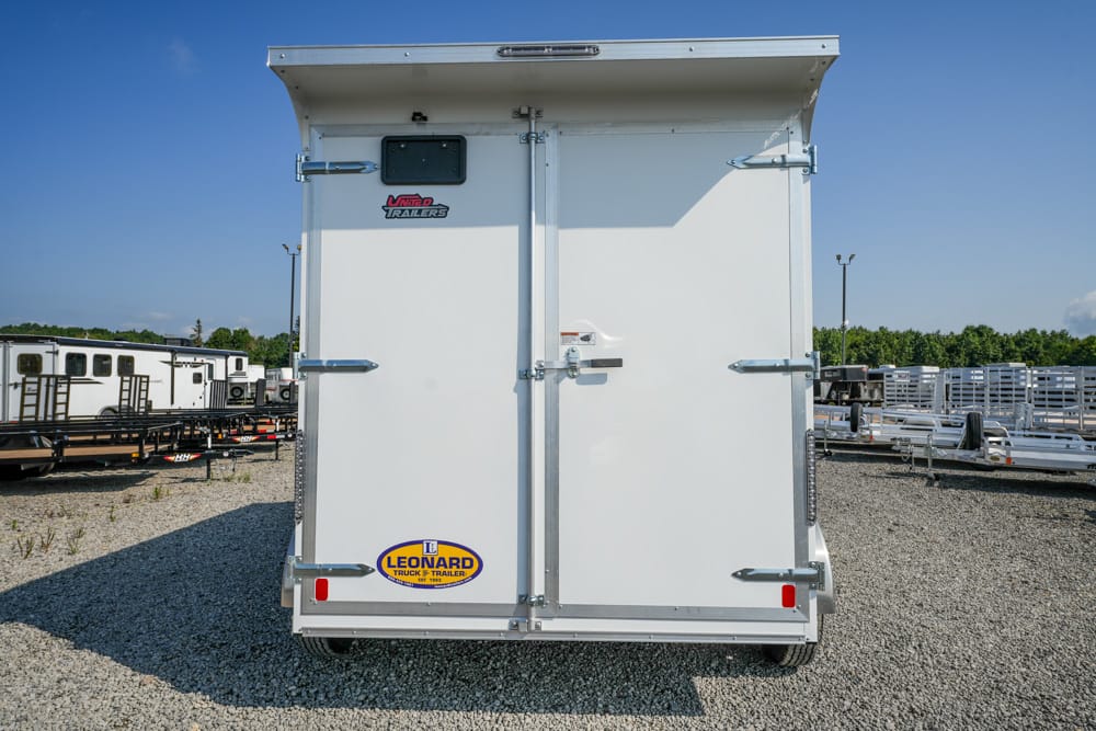 Rear view of a white enclosed cargo trailer with dual back doors, metal latches, and a Leonard trailer sticker on a gravel lot.