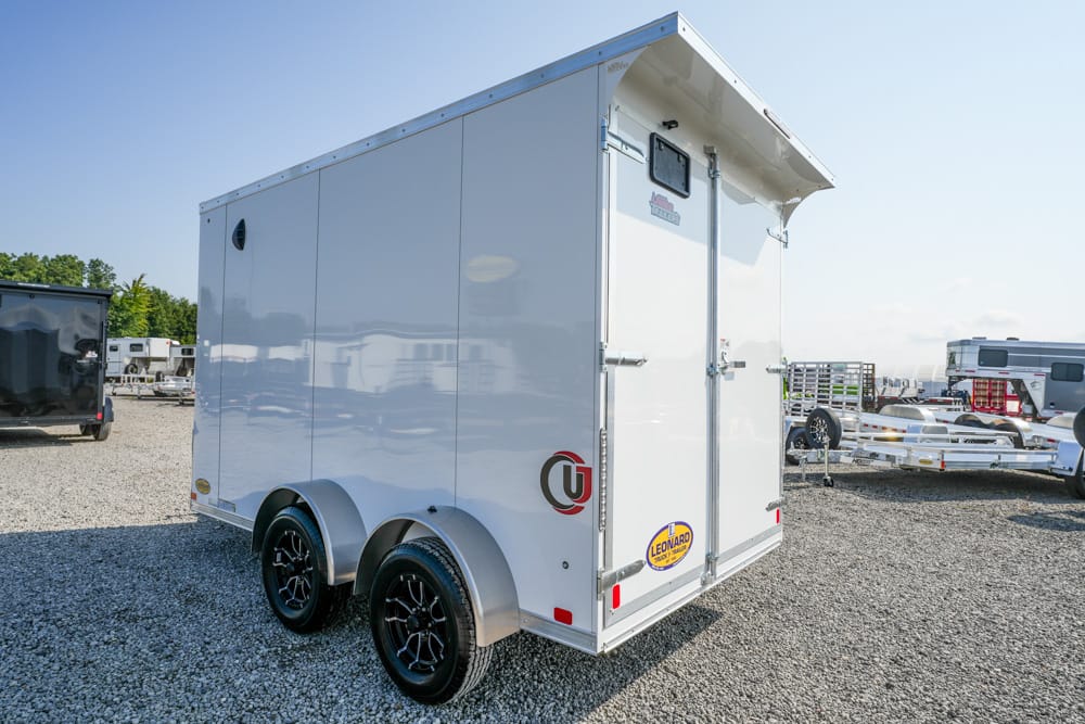 White enclosed cargo trailer with two axles and black rims parked on a gravel lot.