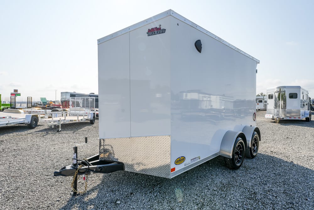 White enclosed dual-axle cargo trailer on a gravel lot, hitch and diamond-plate front, with other trailers in the background.