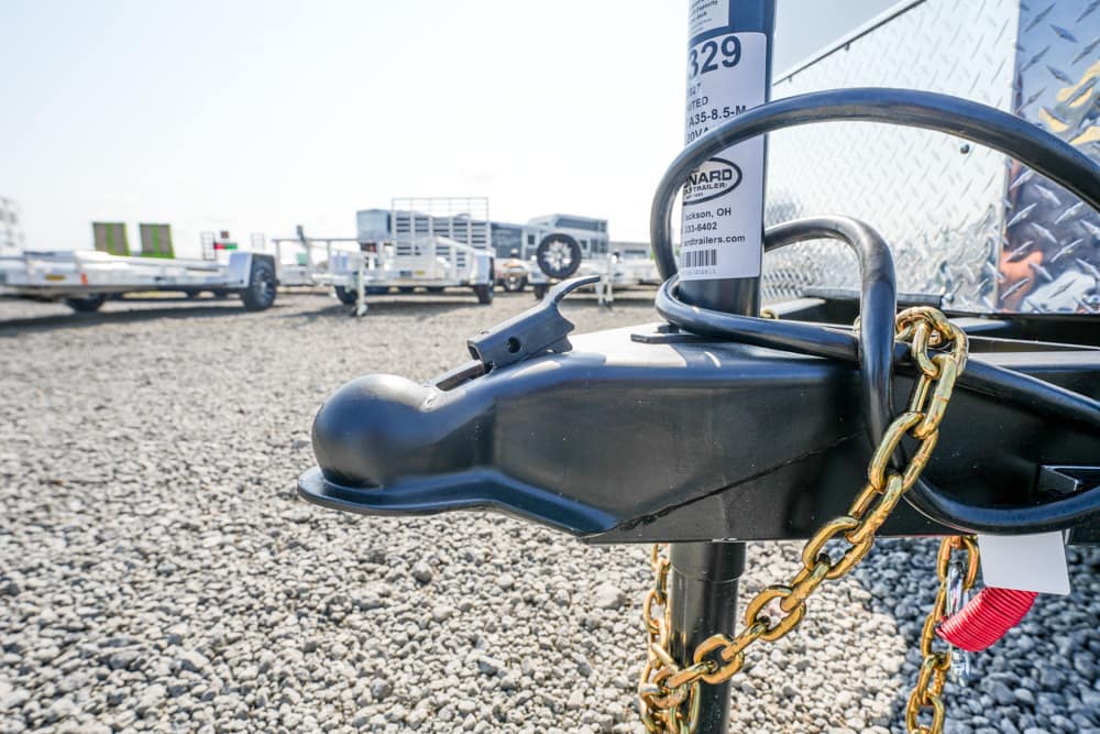 Close-up of a trailer hitch with a gold chain and padlock securing the hitch on a gravel lot.