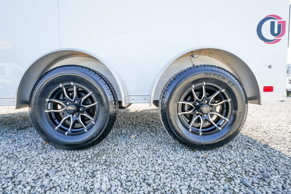 Dual trailer wheels with black alloy rims resting on a gravel surface beneath a white trailer body