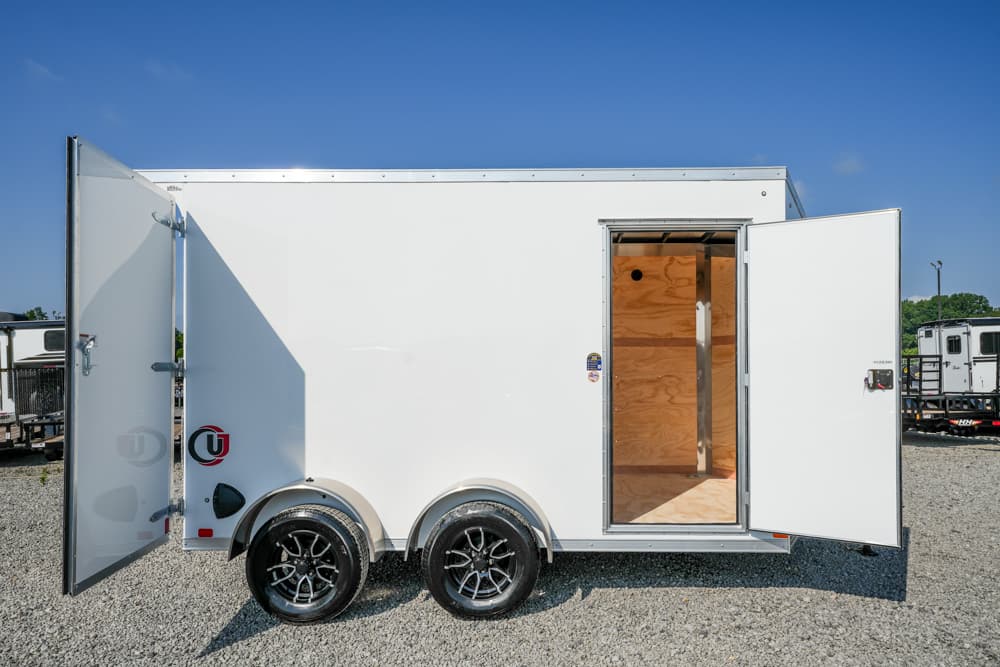 White enclosed cargo trailer with rear and side doors open, showing plywood interior and dual-axle wheels on a gravel lot under a blue sky.