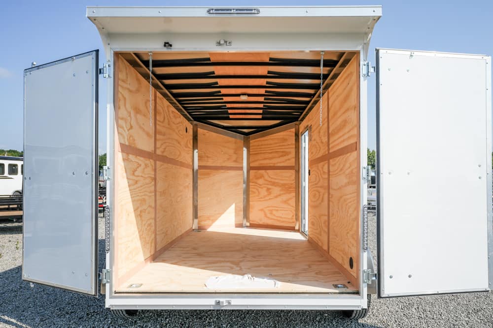 Empty cargo trailer interior with plywood walls and open rear doors, parked on a gravel lot.