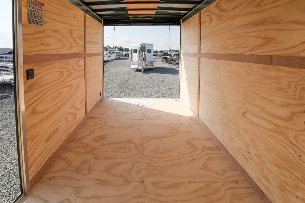 Interior of an empty plywood-walled trailer, open rear door reveals a gravel lot with parked trailers in the distance.