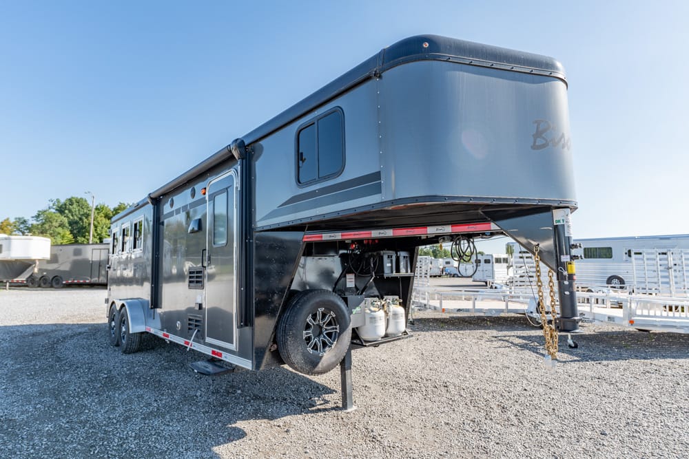 Gooseneck fifth-wheel trailer with living quarters parked on gravel, spare tire and propane tanks underneath.