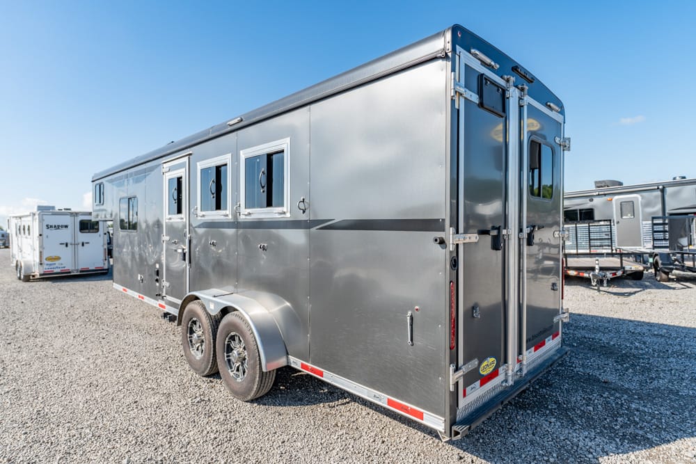 Silver aluminum horse trailer with two axles and multiple small windows, parked on a gravel lot among other trailers at a dealership or storage yard.