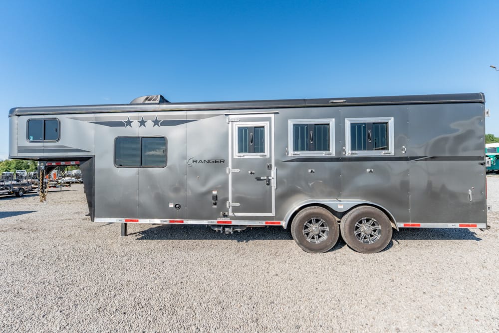Grey horse trailer with a star decal and three side windows, parked on a gravel lot under a clear blue sky.