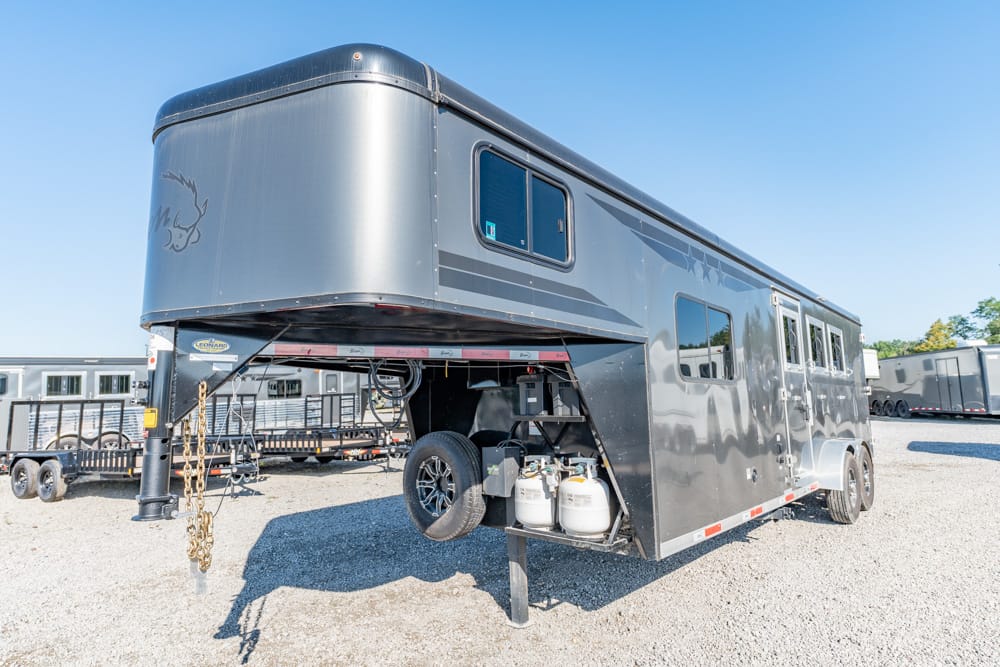 Gray gooseneck horse trailer with living quarters, parked on a gravel lot; propane tanks and a spare tire visible beneath the front section, with windows along the side.