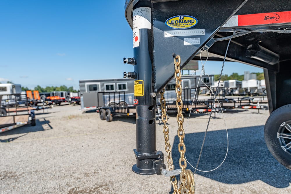 Close-up of a gooseneck trailer hitch with gold safety chains and cables, in a gravel lot with other trailers in the background under a blue sky