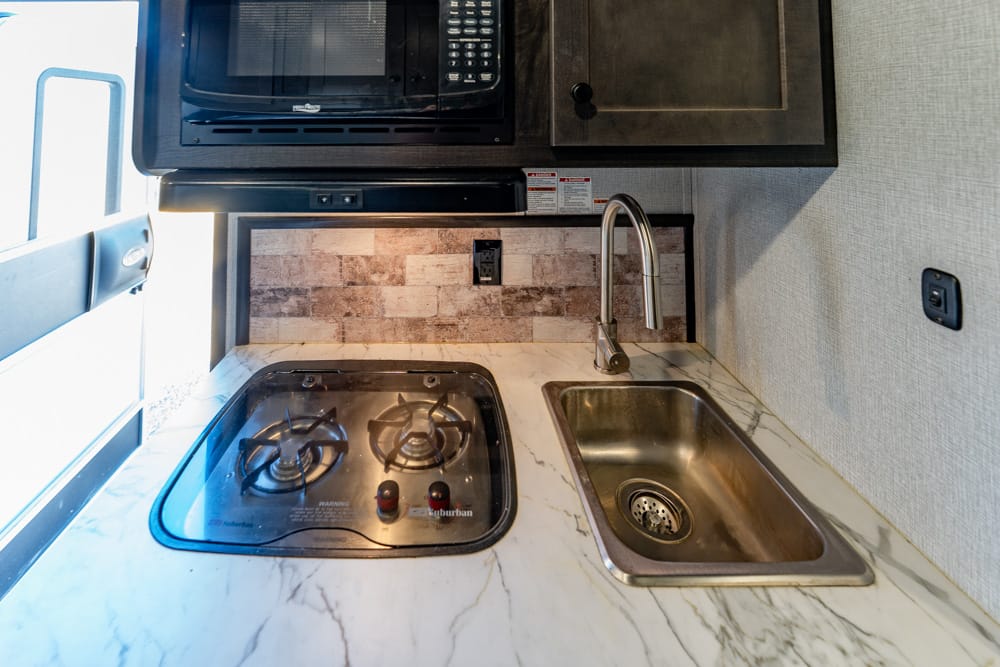 Compact RV kitchenette with a two-burner gas stove, marble countertop, and a stainless steel sink under a mounted microwave.