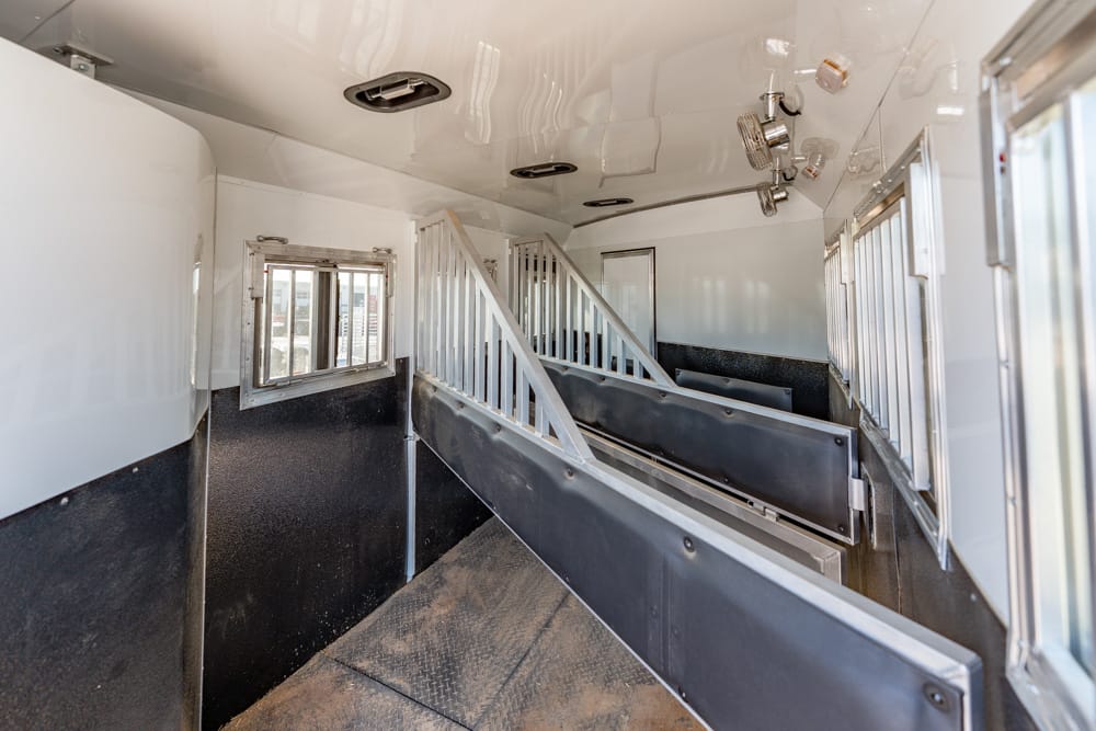 Interior of a white horse trailer with metal stall dividers and small windows, showing a dirt floor and dividing walls for animals in transport.