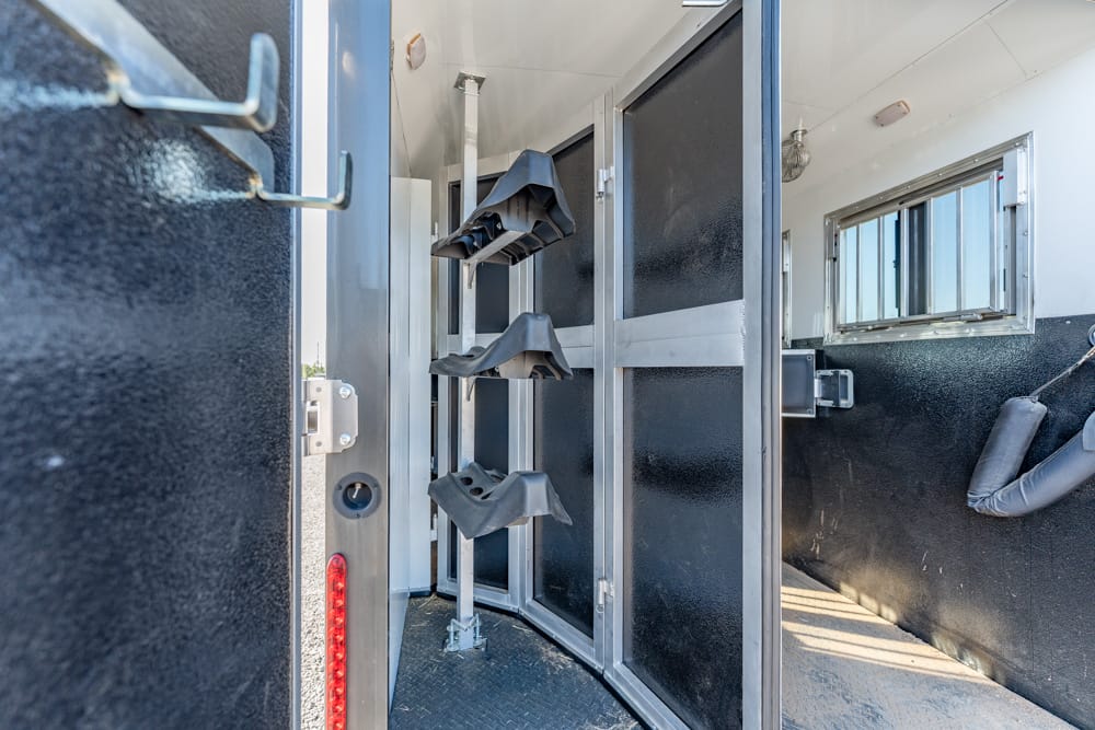 Inside a horse trailer tack room with vertical saddle racks and hanging tack structures along a slim, metal-walled space