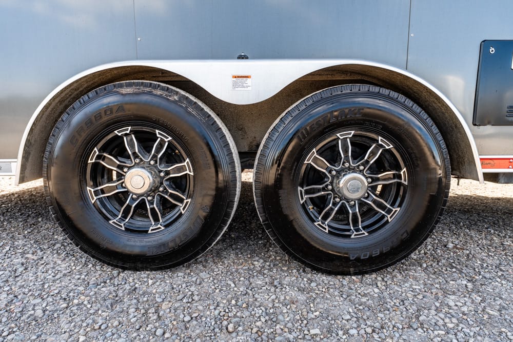 Two black trailer tires with chrome-spoked rims under a silver fender on a gravel surface.
