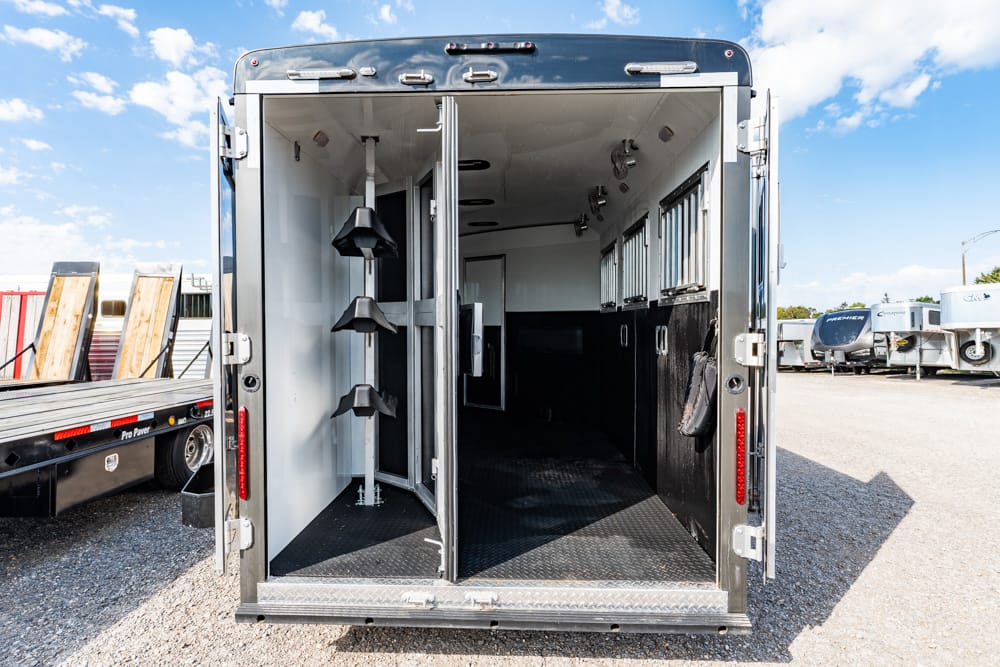 Rear of a black horse trailer with open doors, showing tack storage racks and an empty interior in a gravel lot.