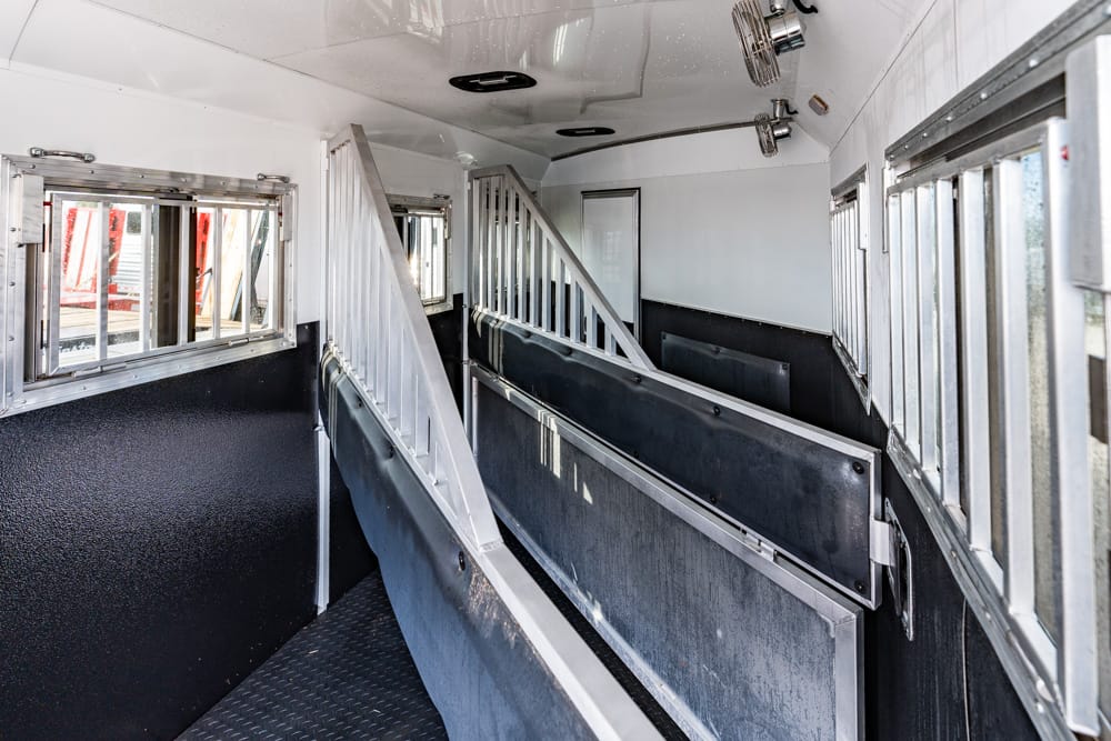 Interior of a horse trailer with partitioned metal stalls and non-slip floor, black lower walls, and windows on the sides