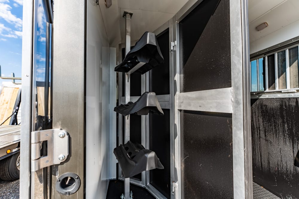 Interior of a metal trailer with vertical saddle racks and black saddle cradles mounted on rails, near an open door.