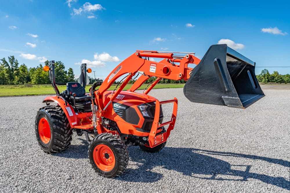 Orange compact tractor with a front-end loader and a large bucket on a gravel lot under a blue sky