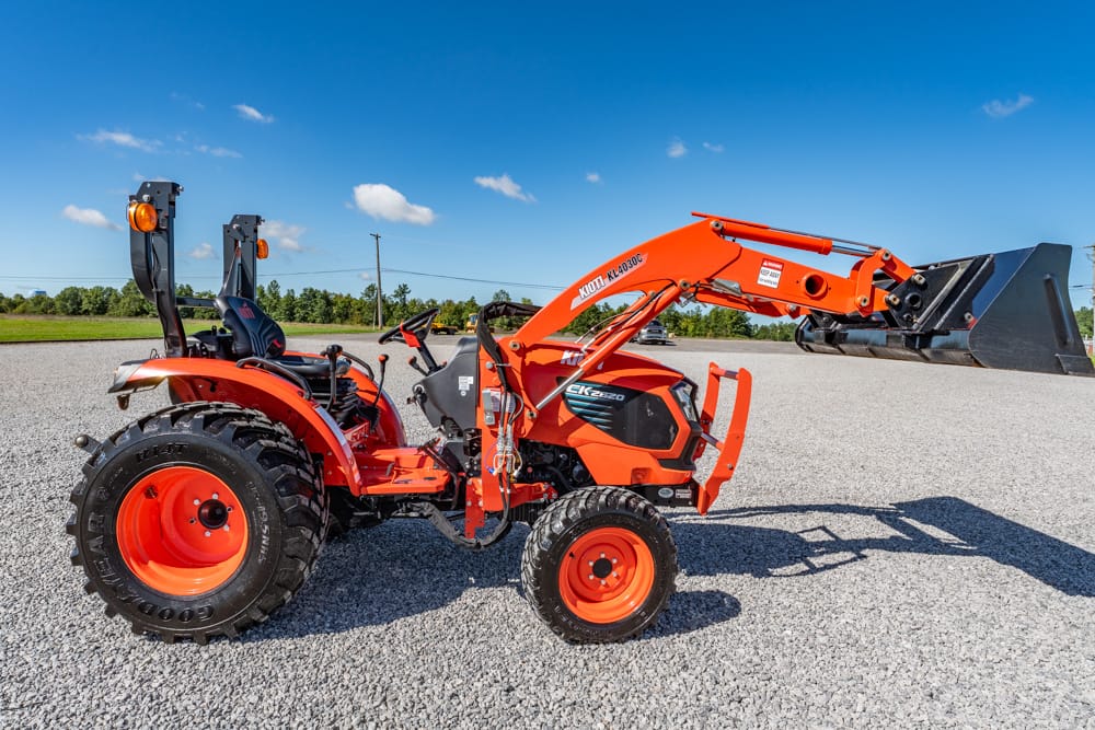 Orange compact tractor with front loader raised, parked on gravel under a blue sky, ready for work.