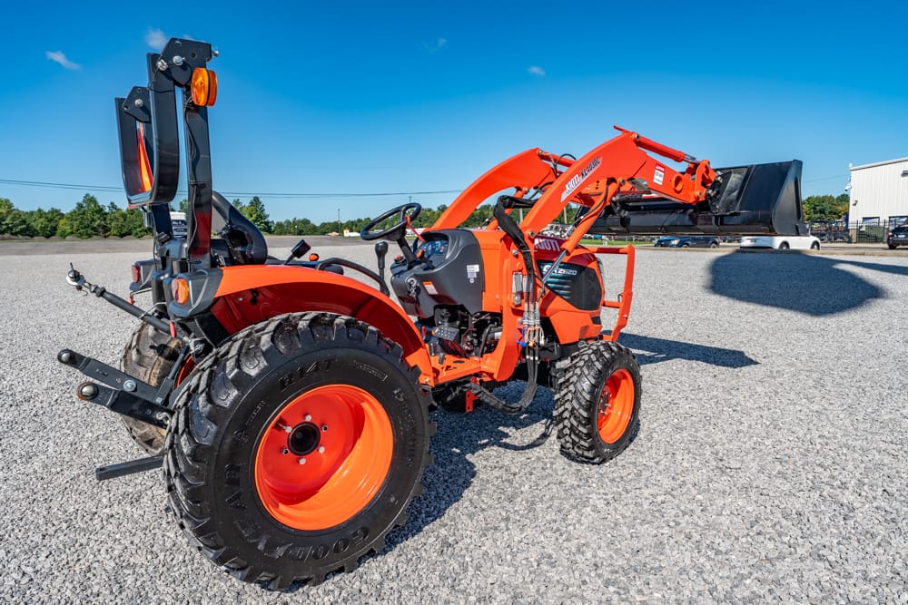 Orange compact tractor with a front loader and rear backhoe parked on a gravel lot under a clear blue sky.