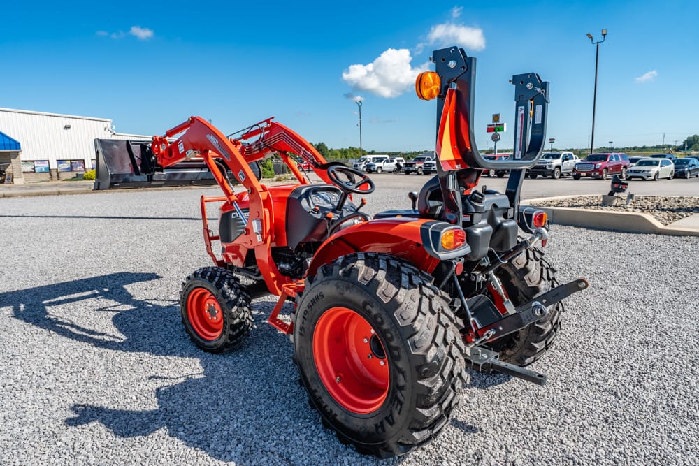 Orange compact tractor with a front loader parked on a gravel lot, blue sky above, dealership in the background.