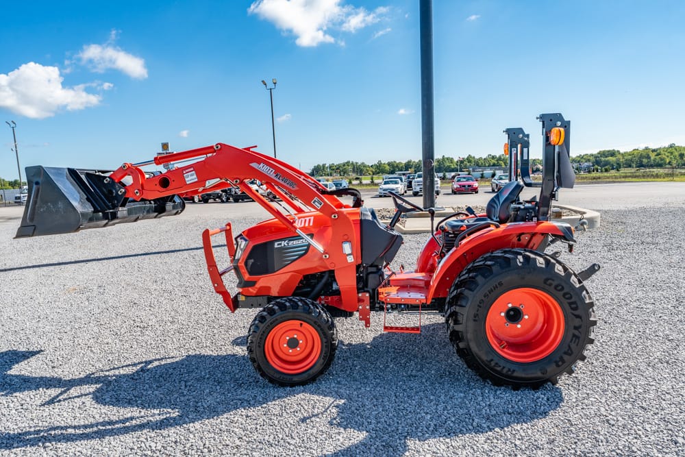 Orange compact tractor with a front-end loader raised, parked on a gravel lot under a blue sky.