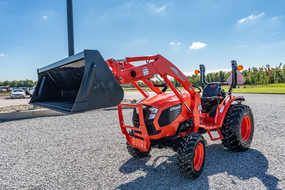 Orange compact utility tractor with a large front-end loader bucket parked on gravel lot, clear blue sky above.