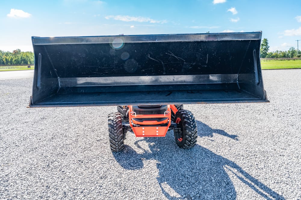 Orange compact loader with a raised steel bucket on a gravel lot under a bright blue sky.