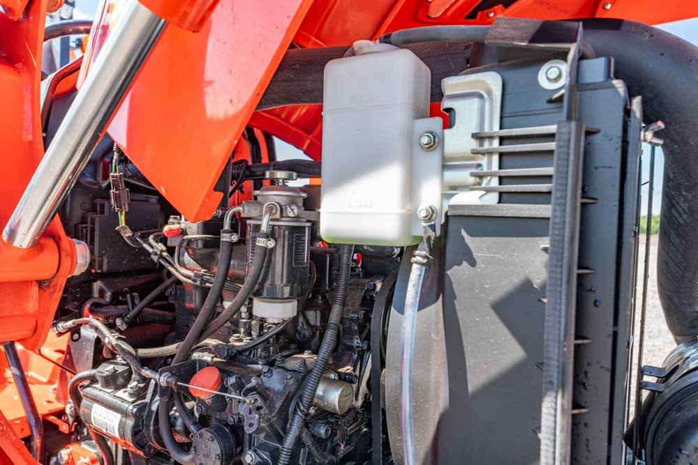 Close-up of a red tractor engine showing hoses, a white coolant reservoir, and a radiator core with metal components.
