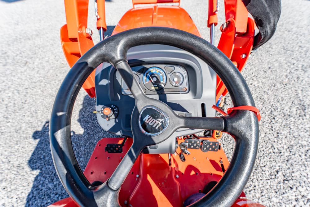 Top-down view of a red tractor cockpit with a black steering wheel and gray dashboard on a gravel surface.