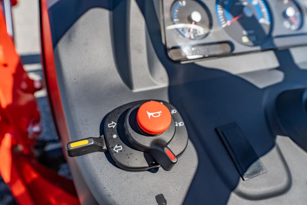 Red horn button on a joystick-style control mounted to a gray vehicle dashboard, with instrument gauges blurred in the background and a yellow marker on a nearby lever.