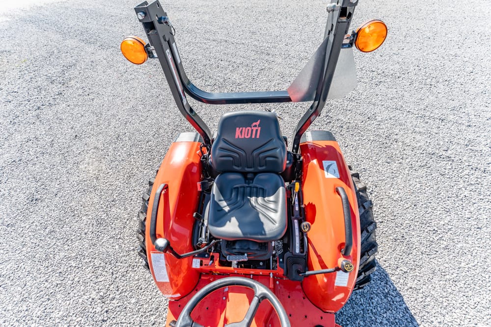 Overhead view of an orange Kioti compact tractor, showing the black seat, roll bar, and large tires on a gravel surface.