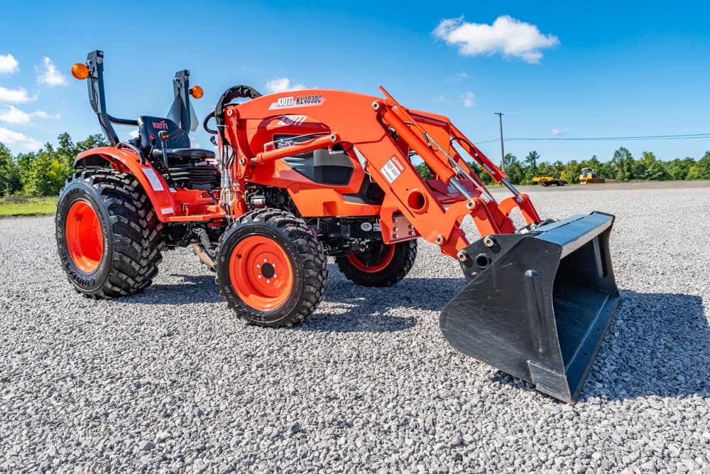 Orange compact tractor with a hydraulic front loader parked on a gravel lot under a clear blue sky.