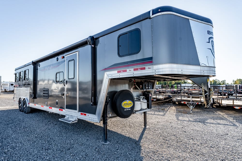 Large silver gooseneck RV trailer parked on gravel lot, front hitch visible with spare tire mounted under the nose.