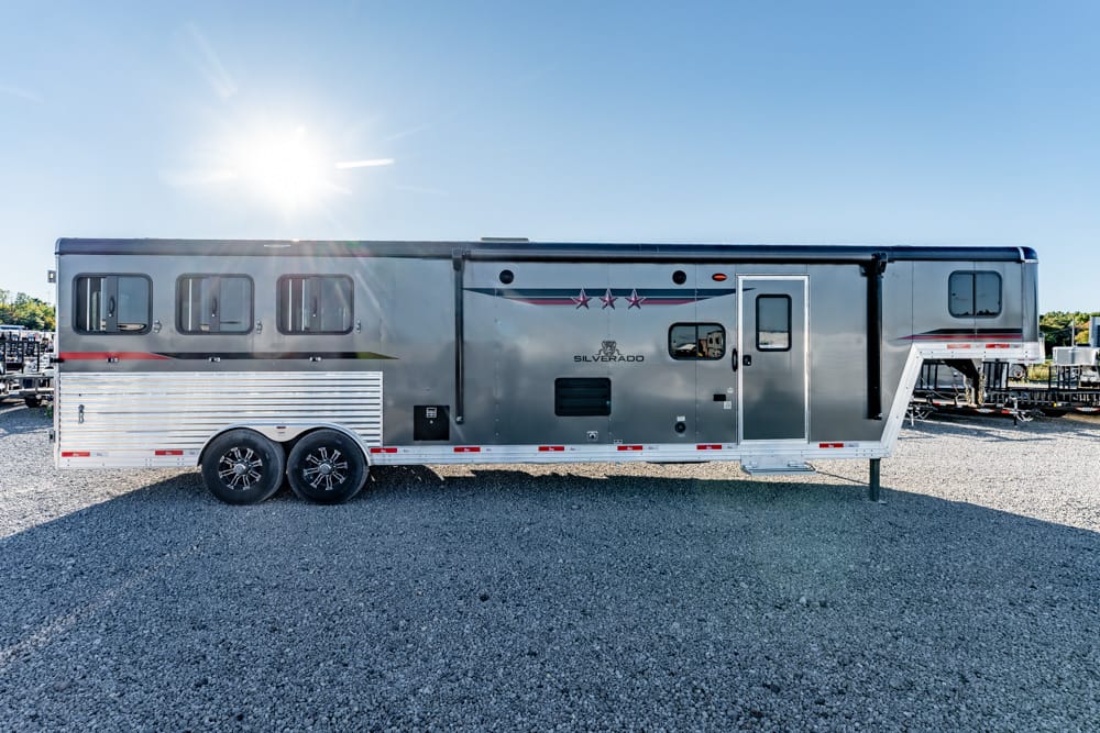 Long silver fifth-wheel trailer with double axles parked on a gravel lot under a bright sun.